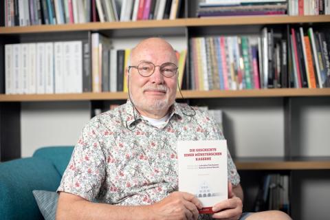Ein Mann sitzt auf einem blauen Sofa vor einer Bücherwand und hält ein Buch in Händen