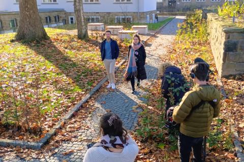 Menschen gehen einen Pfad entlang und werden dabei gefilmt. Herbstlaub liegt auf dem Boden.